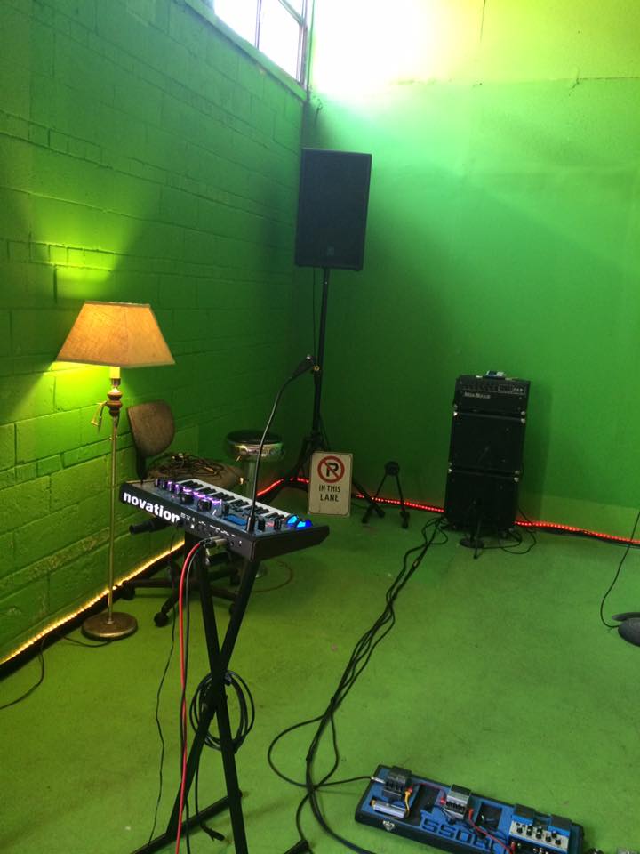Synth on a stand beside a lamp, with pedals and amps on the green floor