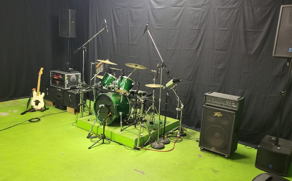 The live room with a green drum kit on a riser, amps and a guitar